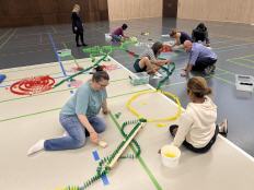 Teilnehmer einer Firmen-Teamchallenge bauen gemeinsam ein buntes Logo aus tausenden Dominosteinen in einer Eventhalle